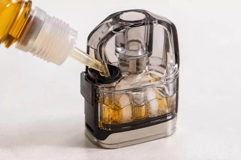 A clear vape pod being filled with e-liquid through a side rubber port from a precision needle tip bottle nozzle.