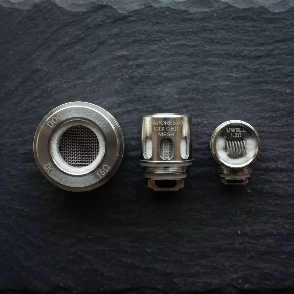 Comparison shot of three different vape coils (0.15 ohm, 0.6 ohm, 1.2 ohm) on a dark slate surface, illustrating physical size difference.