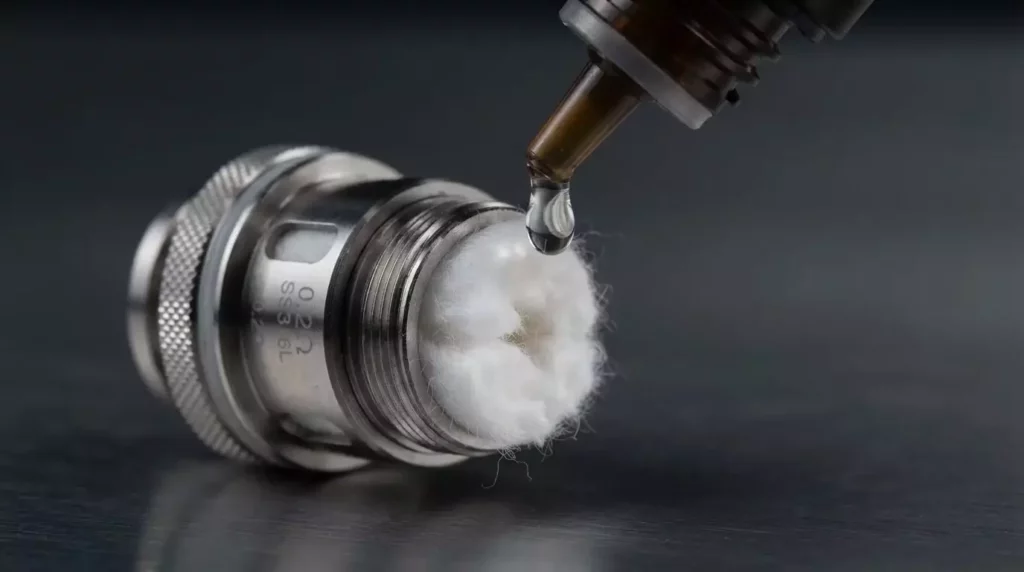 A close-up macro view demonstrating thick high-VG e-liquid being applied to the large cotton wicking port of a sub-ohm vape coil to prevent dry hits.